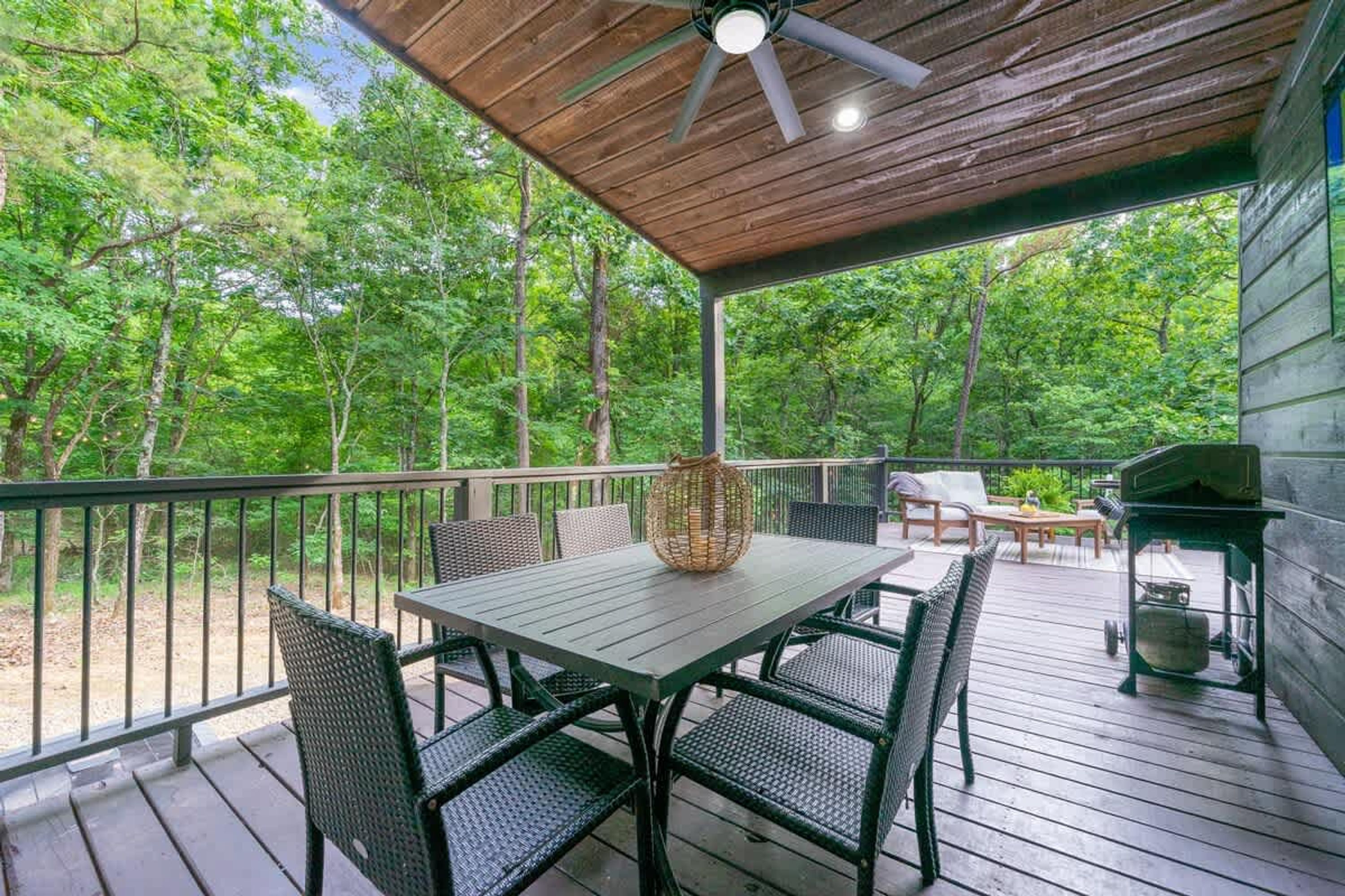 Une terrasse extérieure couverte, dotée d’une table et de chaises, et entourée d’une balustrade en bois et d’arbres.