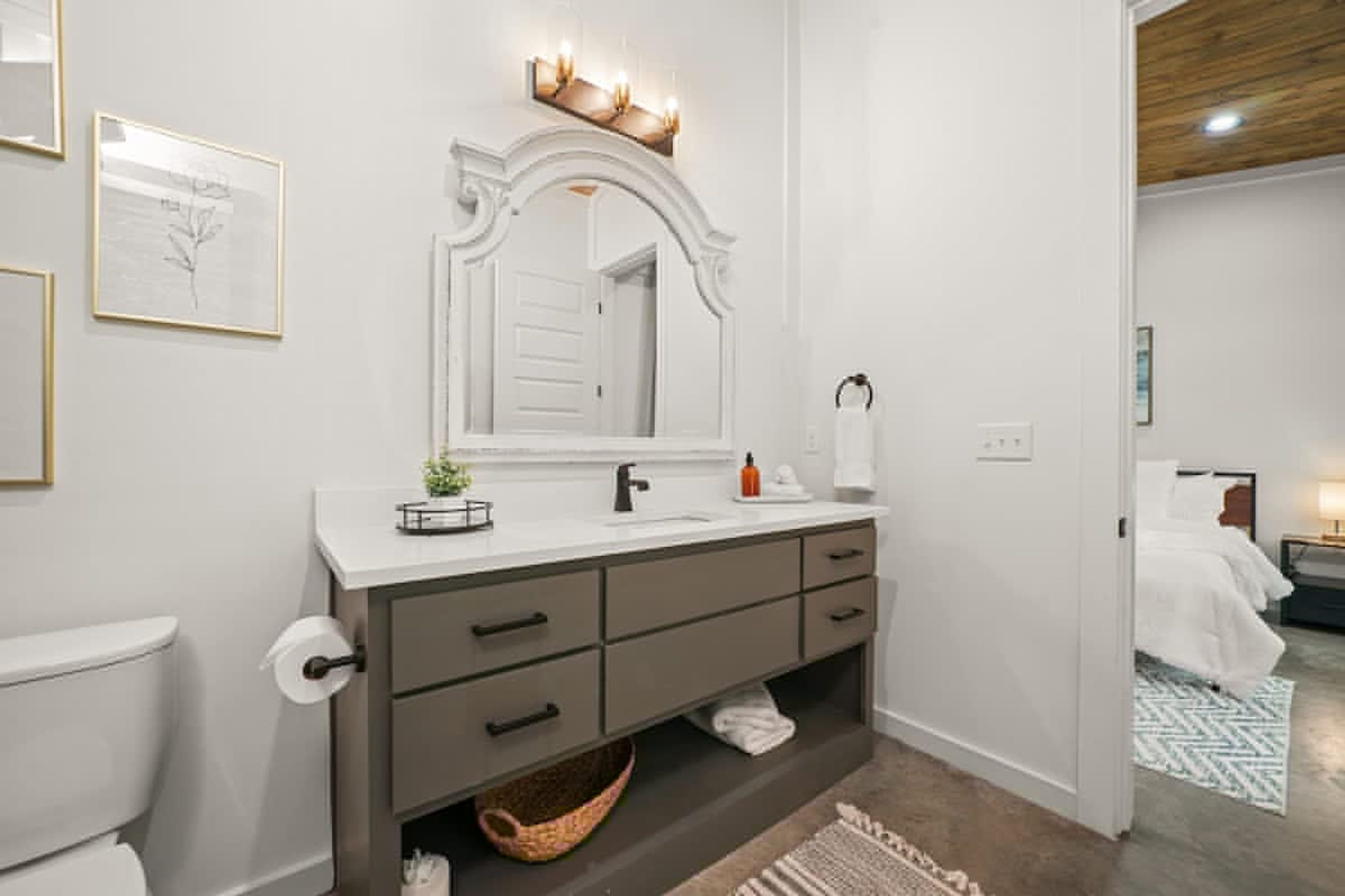 A modern bathroom with a white vanity, dark drawers, a large mirror, and a small basket under the sink.