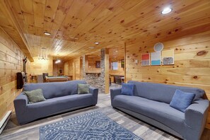 A cozy cabin interior with two sofas, a patterned rug, wooden walls, and a billiards table.