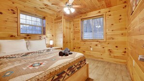Un dormitorio de cabaña de madera con una cama, dos ventanas, un ventilador de techo y una colcha.