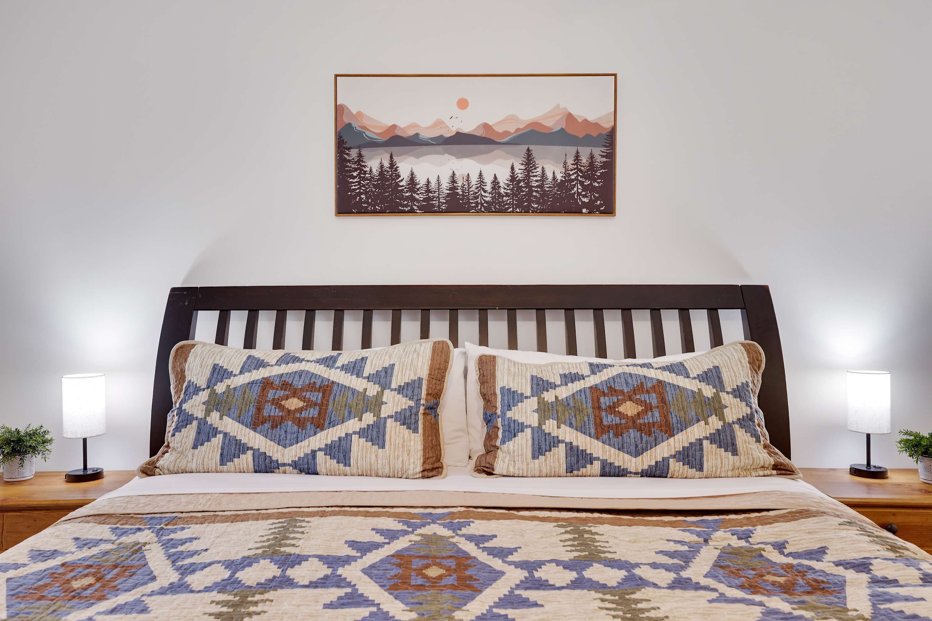 Ein ordentlich bezogenes Bett mit gemusterten Kissen und passender Tagesdecke, ein gerahmtes Landschaftsbild darüber und zwei Nachttische mit Holzoberfläche und je einer Nachttischlampe.