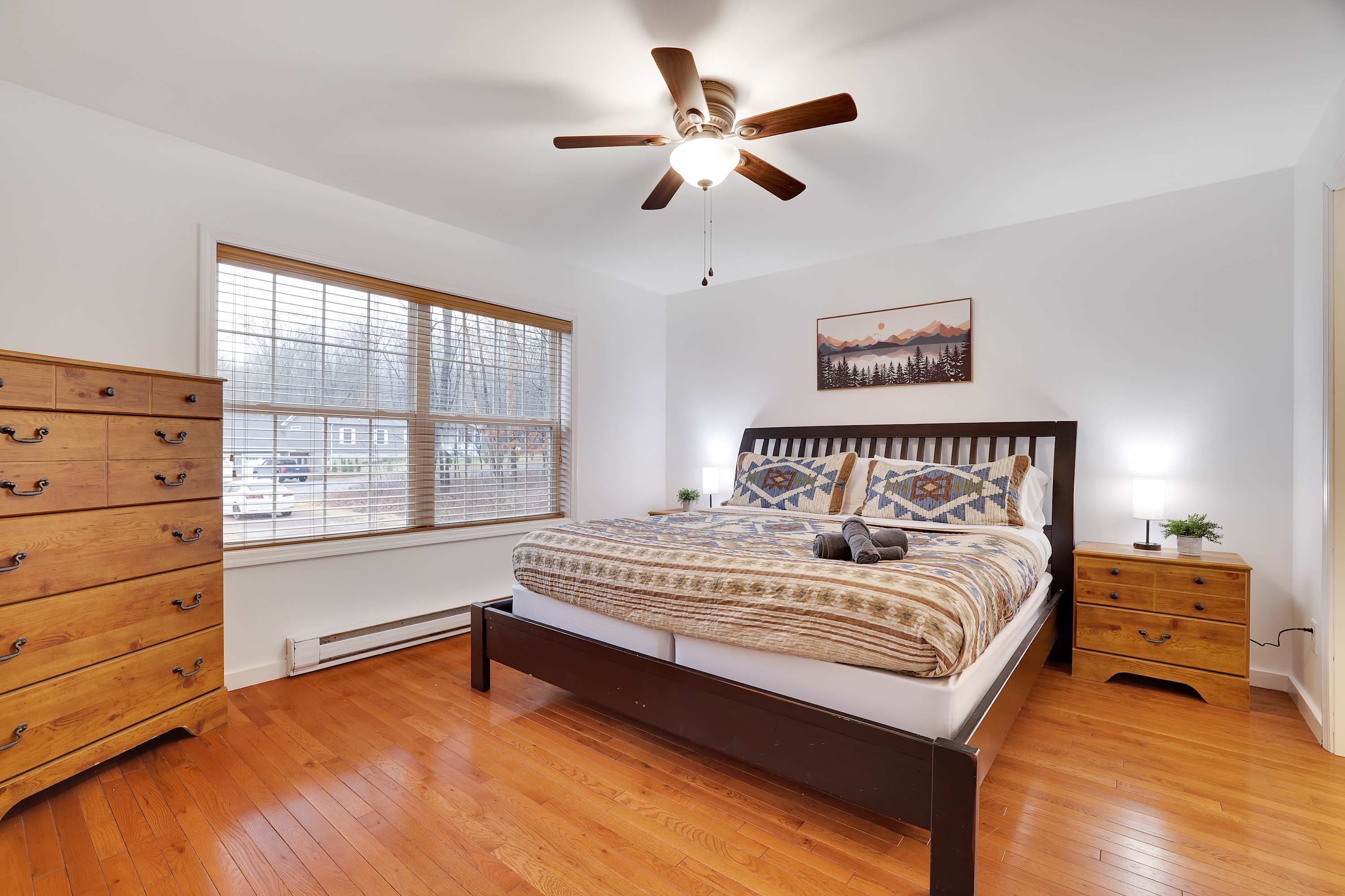 A bedroom with a wooden bed, a nightstand, a dresser, a ceiling fan, and a window with blinds.