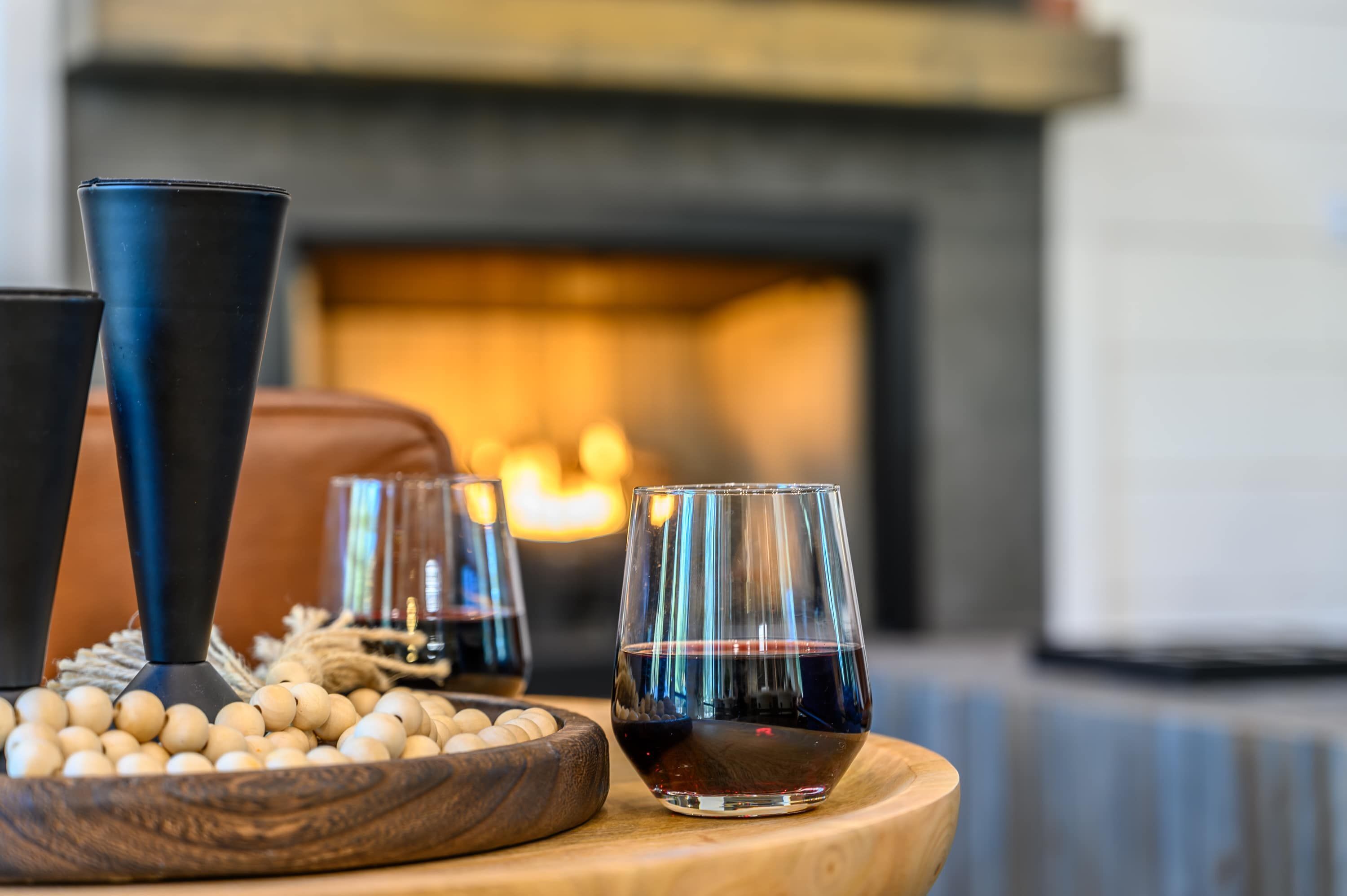 Un ambiente acogedor con chimenea, un cuenco de madera con guijarros blancos y dos copas de vino tinto sobre una mesa de madera.