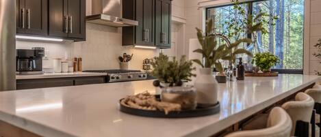 A modern kitchen with dark cabinets, a large island, and a view of a forest through a window.