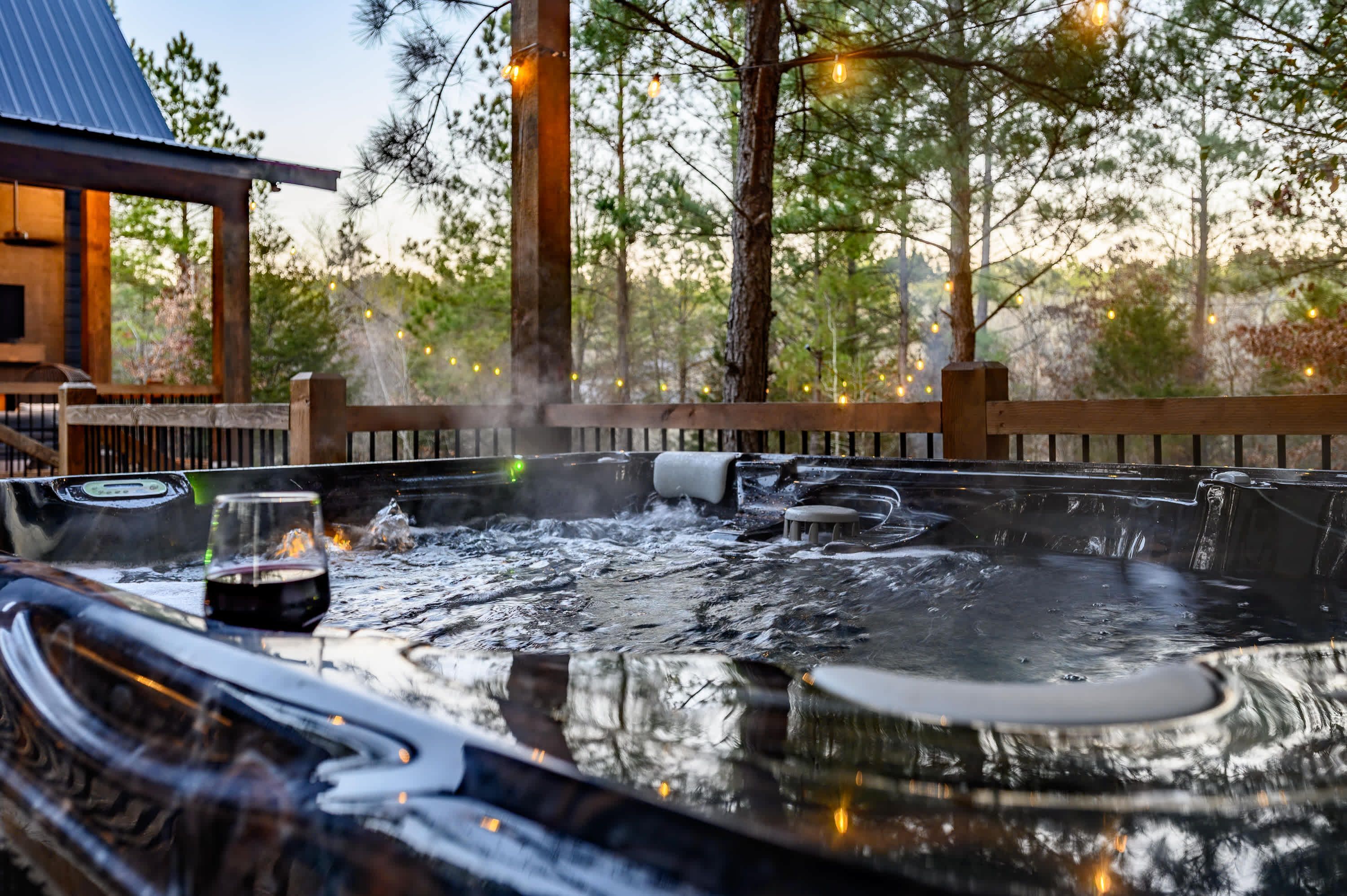 A hot tub with a wooden deck, surrounded by trees and string lights.
