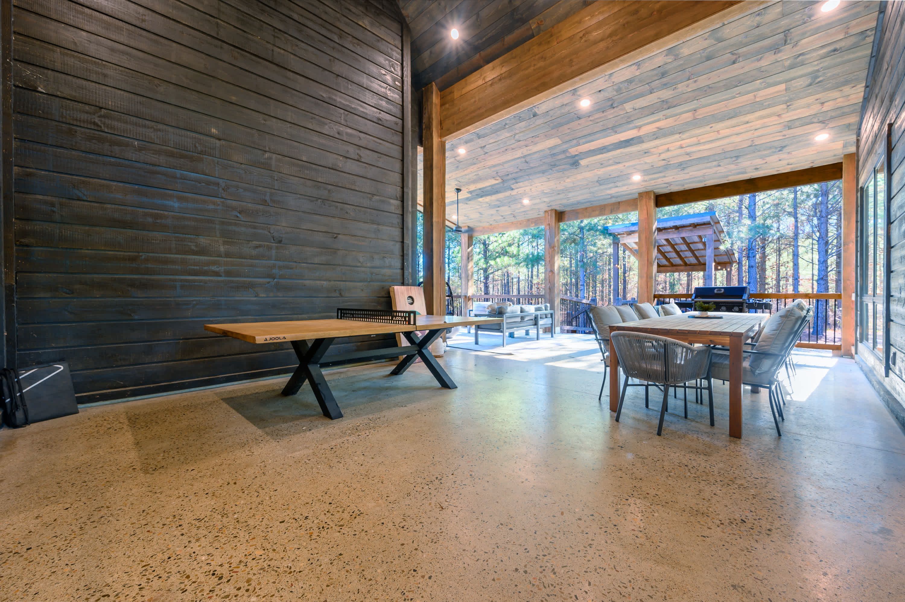 Una habitación amplia con un banco y una mesa de madera, ventanas grandes con vistas al bosque, y una parrilla afuera.