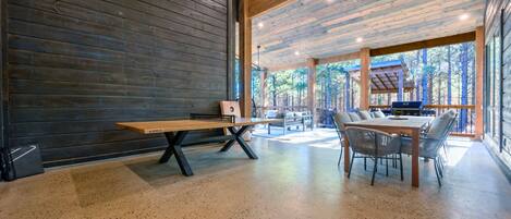 Una habitación amplia con un banco y una mesa de madera, ventanas grandes con vistas al bosque, y una parrilla afuera.