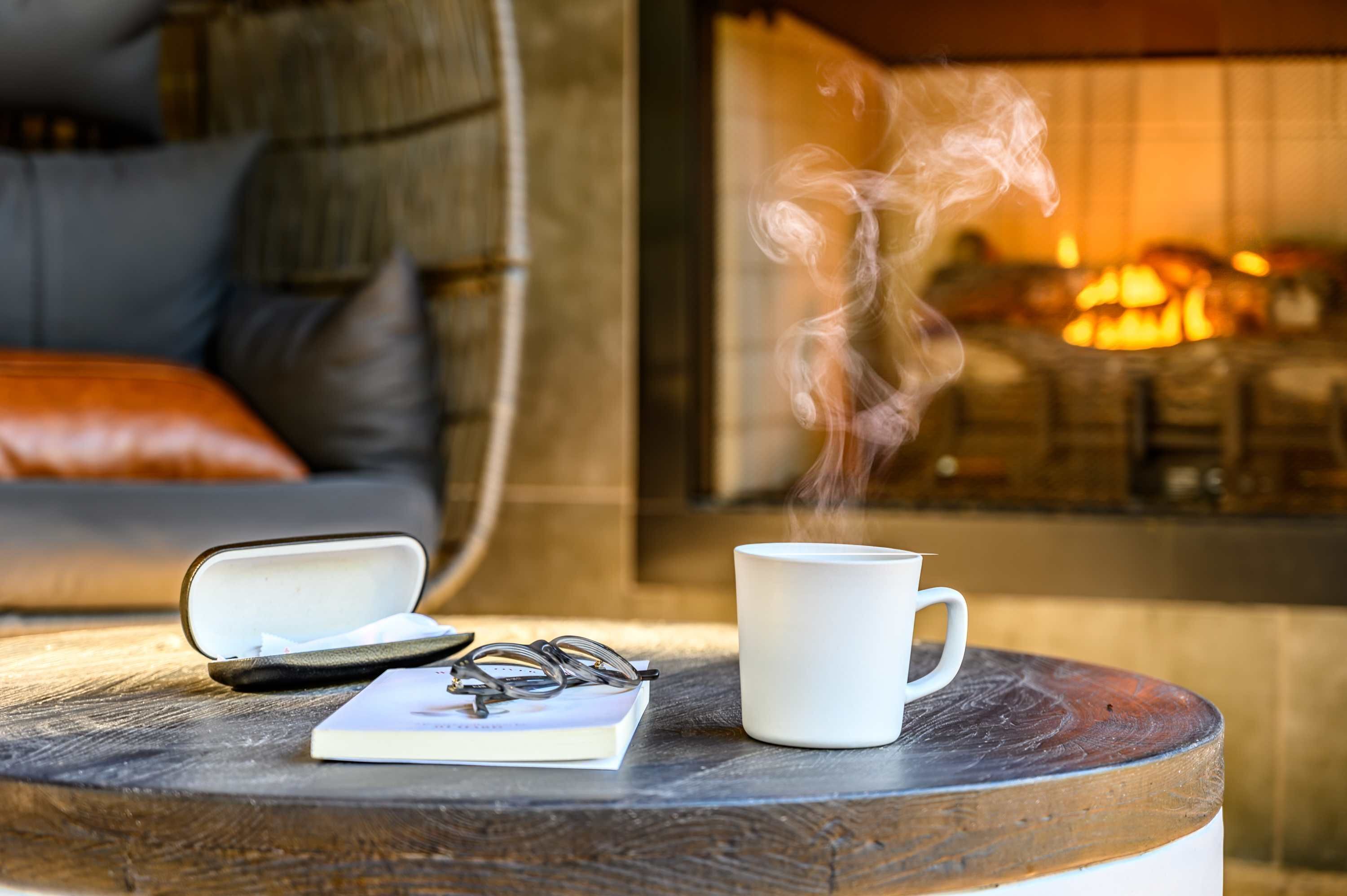 A cozy indoor setting with a fireplace, a round wooden table with a white mug, a pair of glasses, and a notebook.
