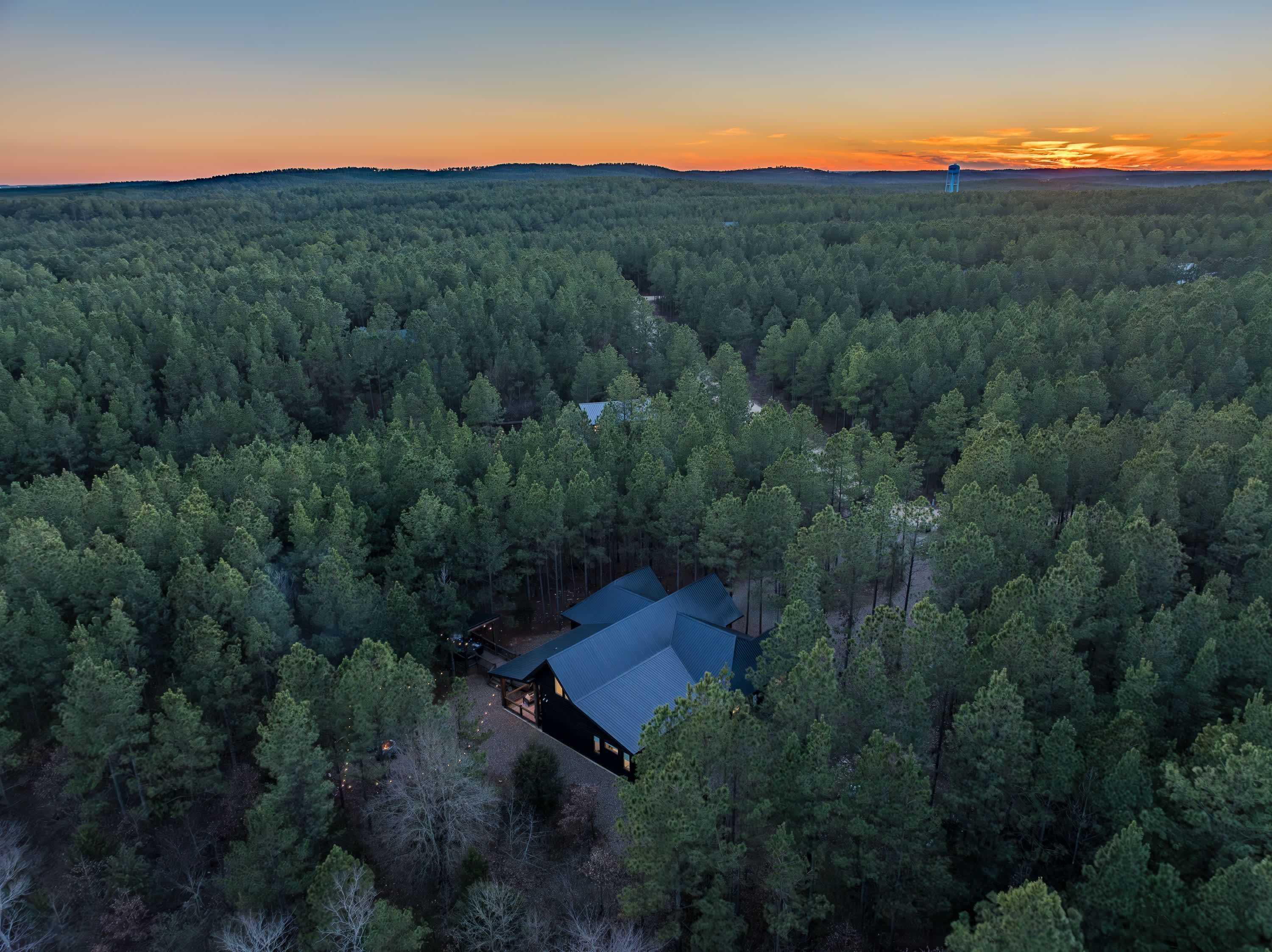 Una cabaña rodeada de bosque denso, con una vista despejada del atardecer al fondo.