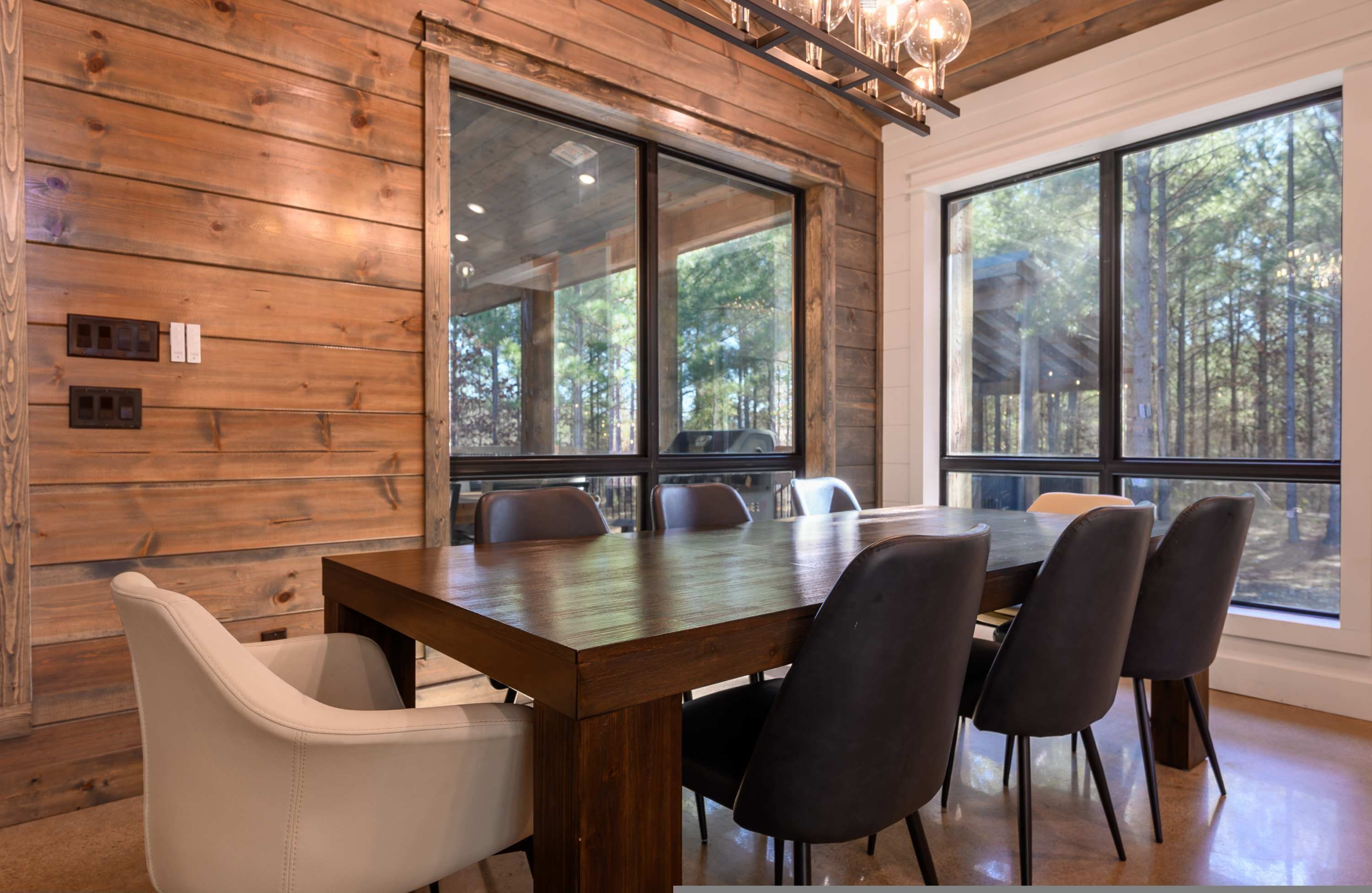 A dining area with a wooden table and chairs, large windows with a forest view, and a chandelier.