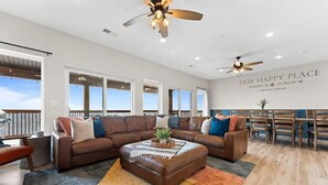 A spacious living room with a large brown sectional sofa, a coffee table, and a ceiling fan. There are large windows providing natural light and a view of the outdoors. - Modern Getaway w Hot Tub Arcade Stunning Views (Branson)