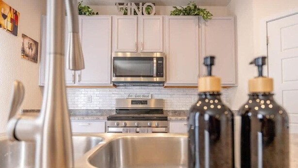 A kitchen with white cabinets, a stainless steel sink, a microwave, and a stove. There are two black bottles with yellow caps on the counter. - Elegant Spacious 4BR Home Peyton (Elbert)