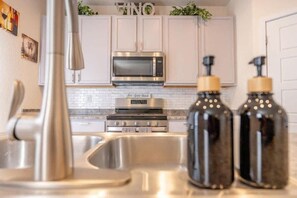 Cocina con armarios blancos, un lavaplatos de acero inoxidable, un microondas y una estufa. Hay dos botellas negras con tapas amarillas sobre la encimera.