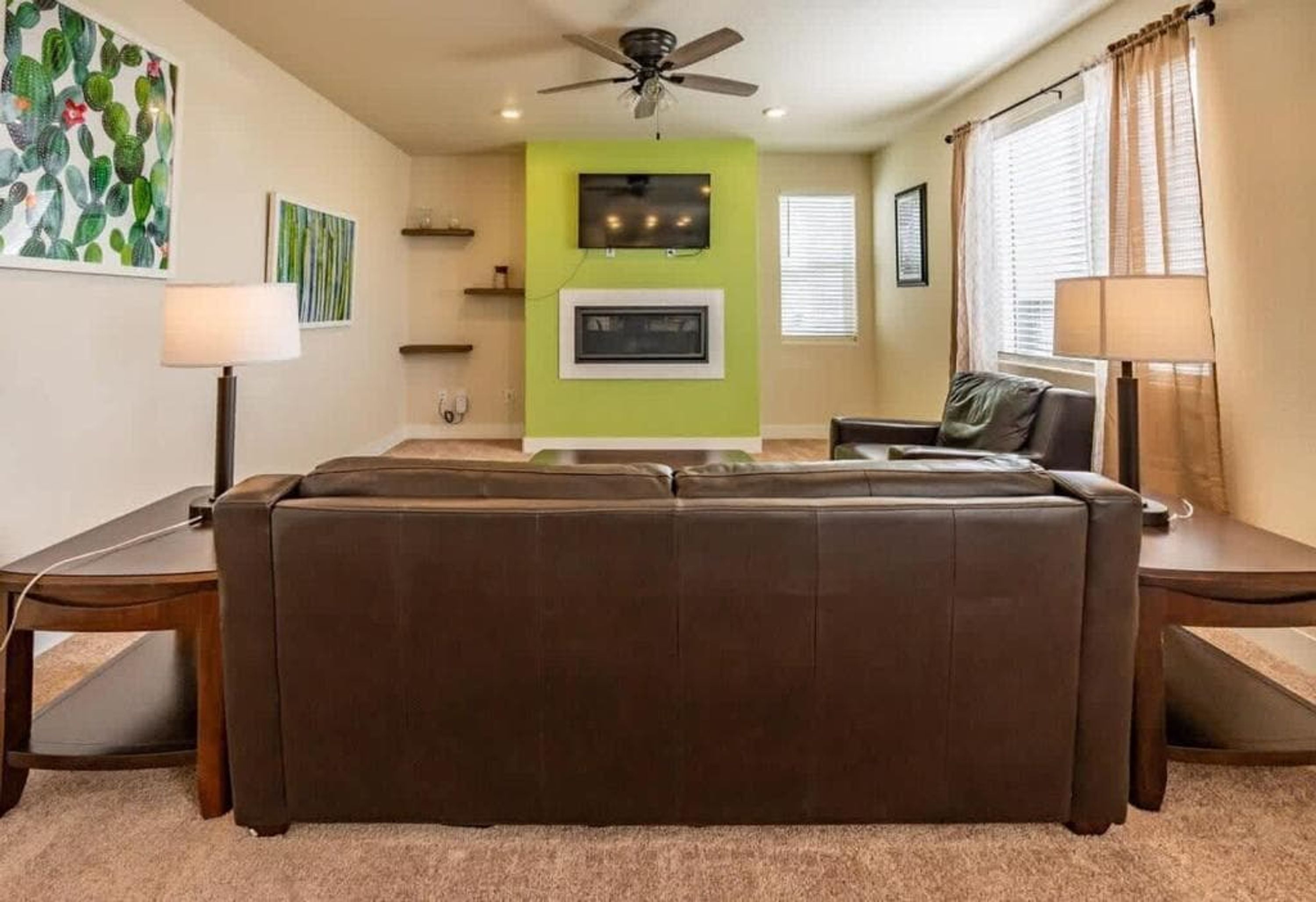 A living room with a brown sofa, a ceiling fan, a TV mounted on the wall, and a window with blinds.