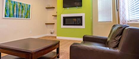 Een woonkamer met een bruine leren bank, een houten salontafel, een aan een groene muur bevestigde televisie en een open haard.