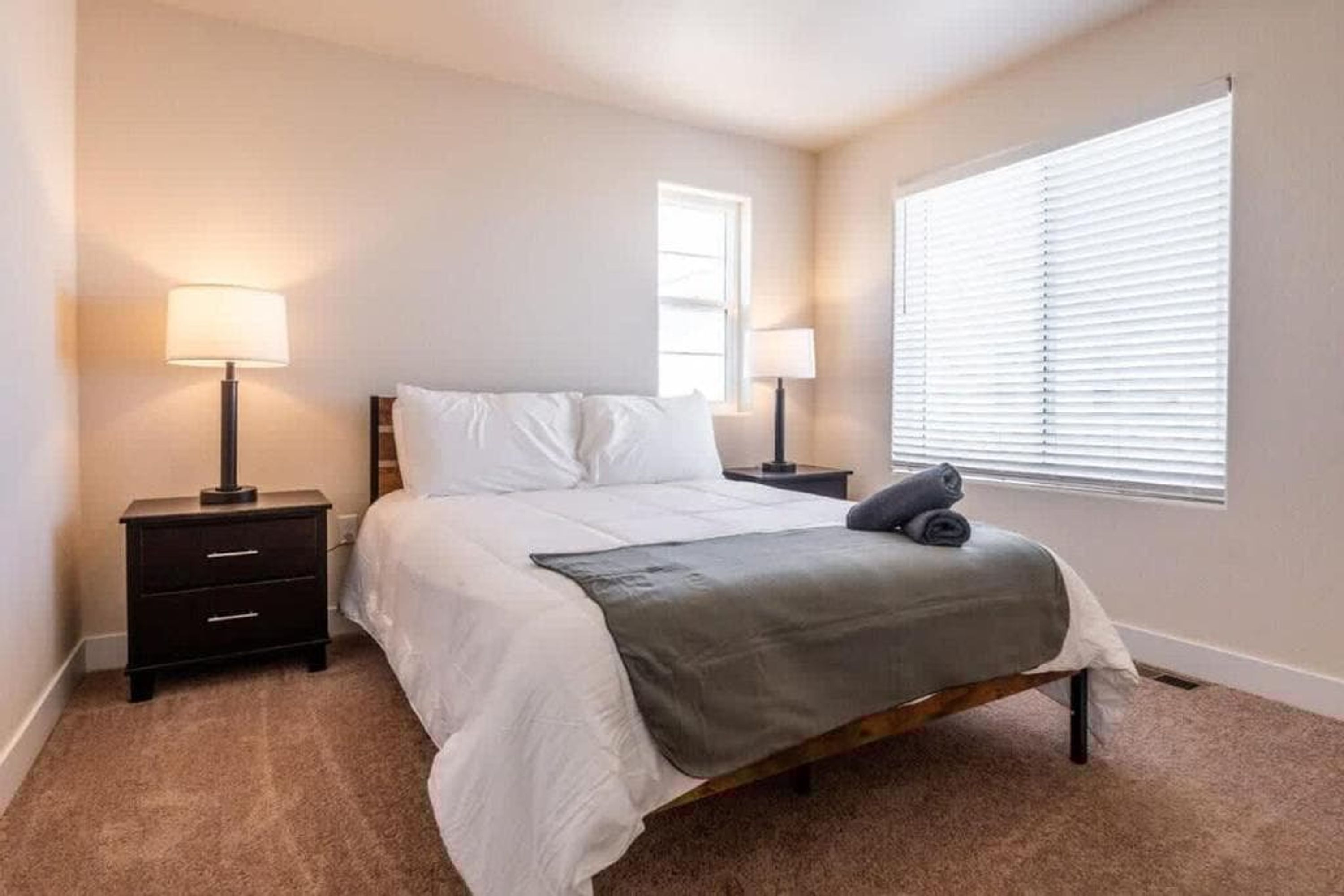 A bedroom with a bed, two bedside tables, a lamp on each table, a window with blinds, and a carpeted floor.