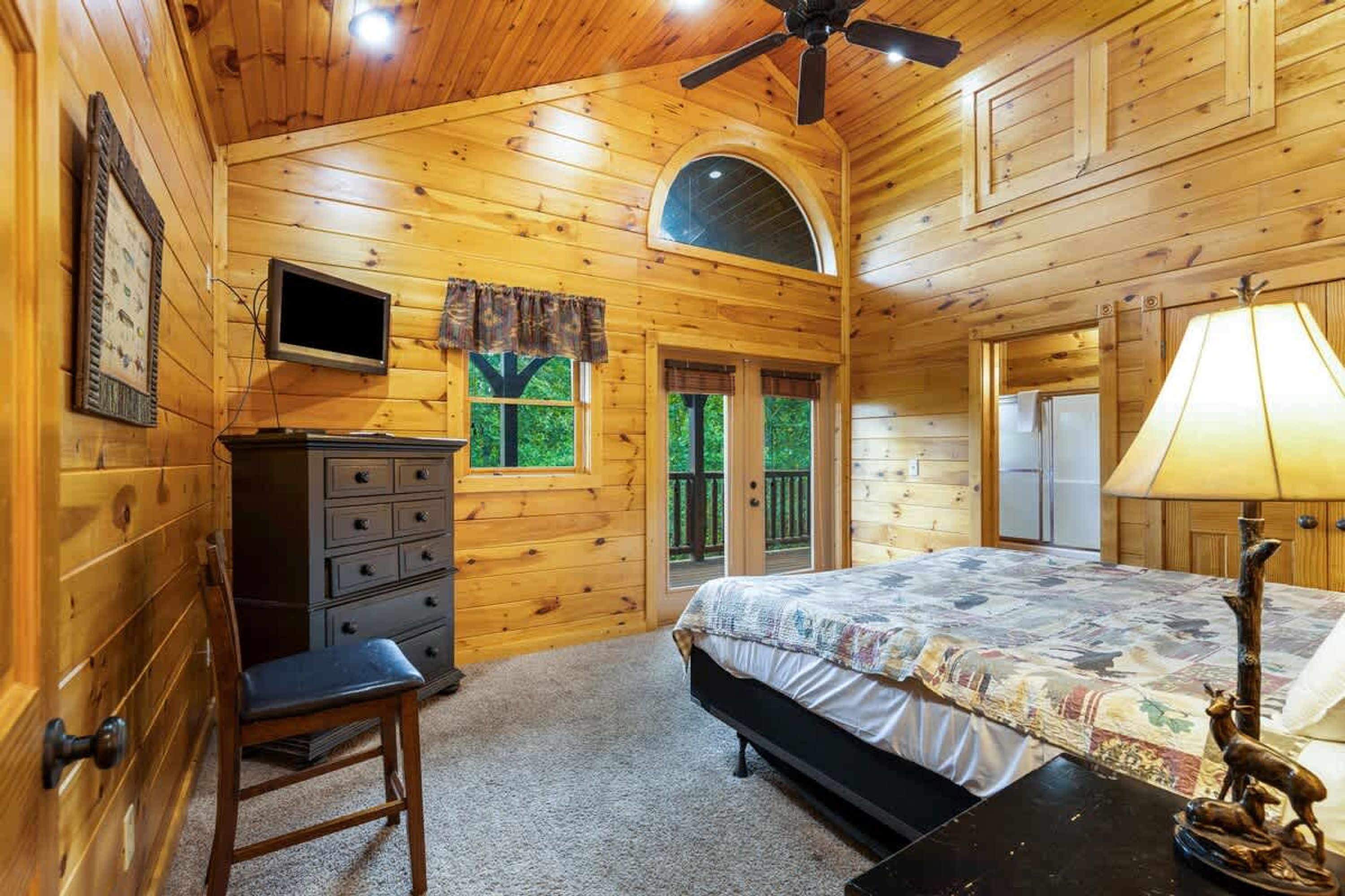 A wooden cabin bedroom with a bed, dresser, chair, TV, and a ceiling fan.