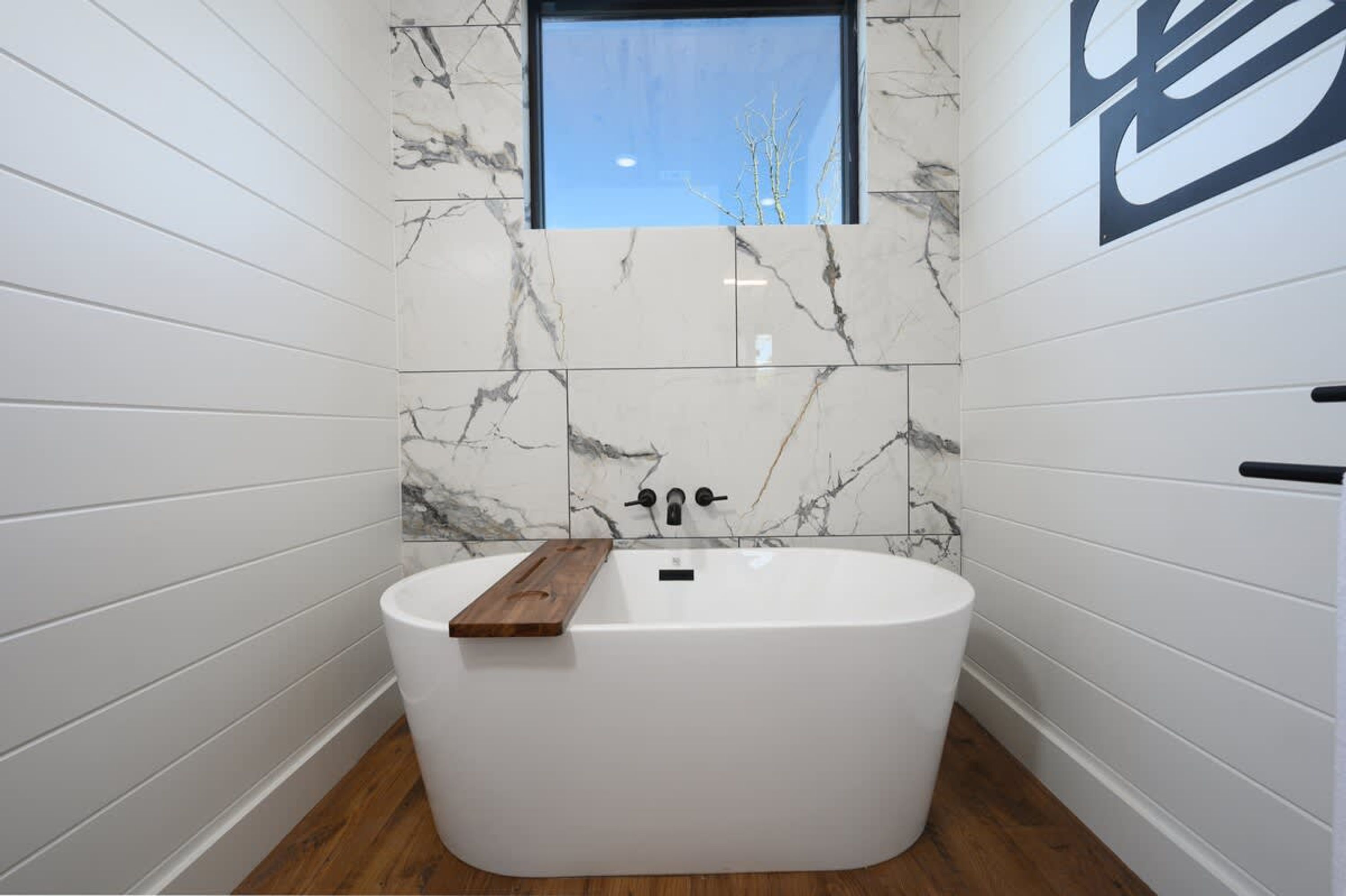 A modern bathroom with a freestanding bathtub, marble wall tiles, and a window above the tub.