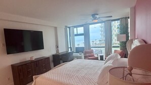 A hotel room with a large bed, a flat-screen TV, a ceiling fan, and a view of the beach through large windows. - Beach Studio With Partial Water Views Sundeck Pool (Daytona Beach Shores)