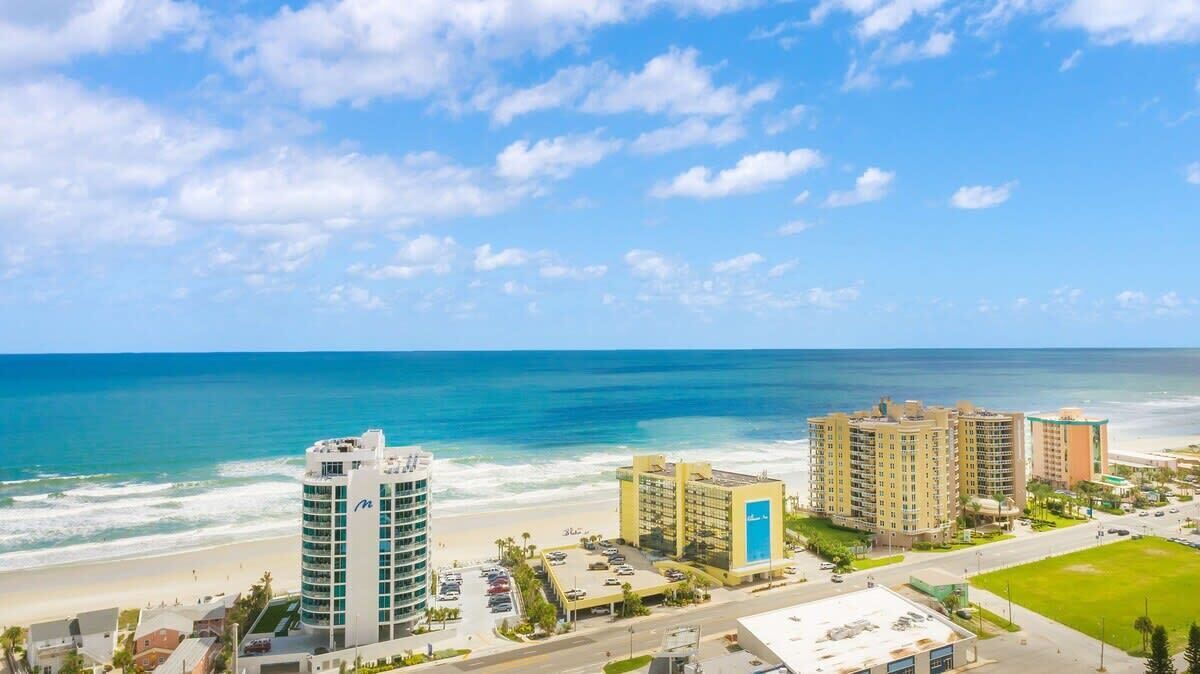 Un hotel frente al mar con varias edificaciones, una playa y un cielo azul despejado.
