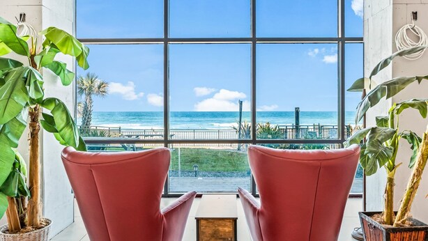 A room with large windows offering a view of the ocean and a pier. There are two red chairs and potted plants. - Beach Studio With Partial Water Views Sundeck Pool (Daytona Beach Shores)