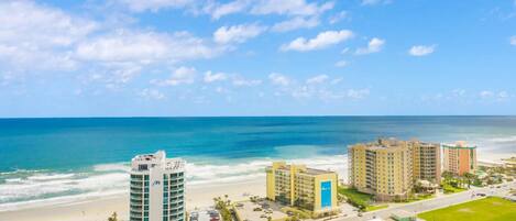A coastal hotel with a beachfront view, featuring a modern building with a blue logo, a parking lot, and a road leading to the beach.