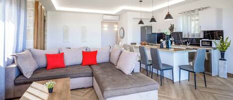 Ein modernes Wohnzimmer mit einem grauen Ecksofa, einem Holztisch und einer Küchenzeile mit Esstisch und Stühlen.