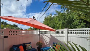 Un espace patio agrémenté d’une clôture blanche en bois, d’un canapé bleu avec des coussins orange et d’un parasol rouge.