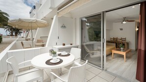 Patio esterno moderno con tavolo rotondo e sedie in rattan bianco, una porta vetrata che dà su un ambiente interno con un letto e una ventola a soffitto, e una palma sullo sfondo.