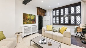A modern living room with a beige sofa, a coffee table with books and a remote, a painting on the wall, and a large window with a view. - Argyl | Phillimore Walk 5 Bed Townhouse (Greater London)
