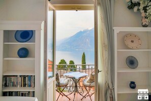 A room with a balcony overlooking a lake and mountains, a table with chairs, and shelves with decorative plates and books. - JULIA'S PLACE by Design Studio (Perledo)