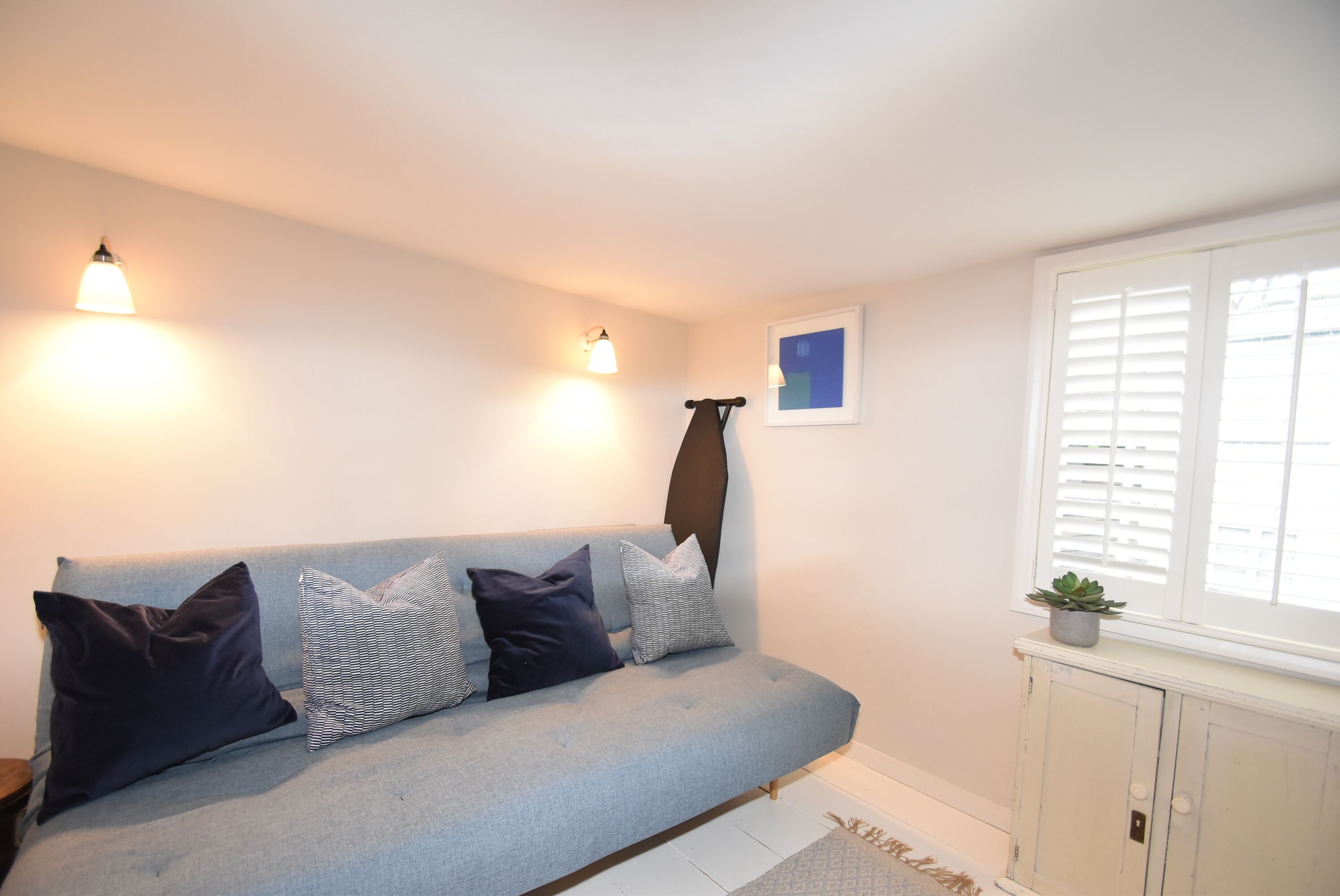 Un canapé bleu clair agrémenté de plusieurs coussins, un luminaire fixé au mur, un tableau encadré, une fenêtre avec des stores et une petite plante posée sur un meuble.