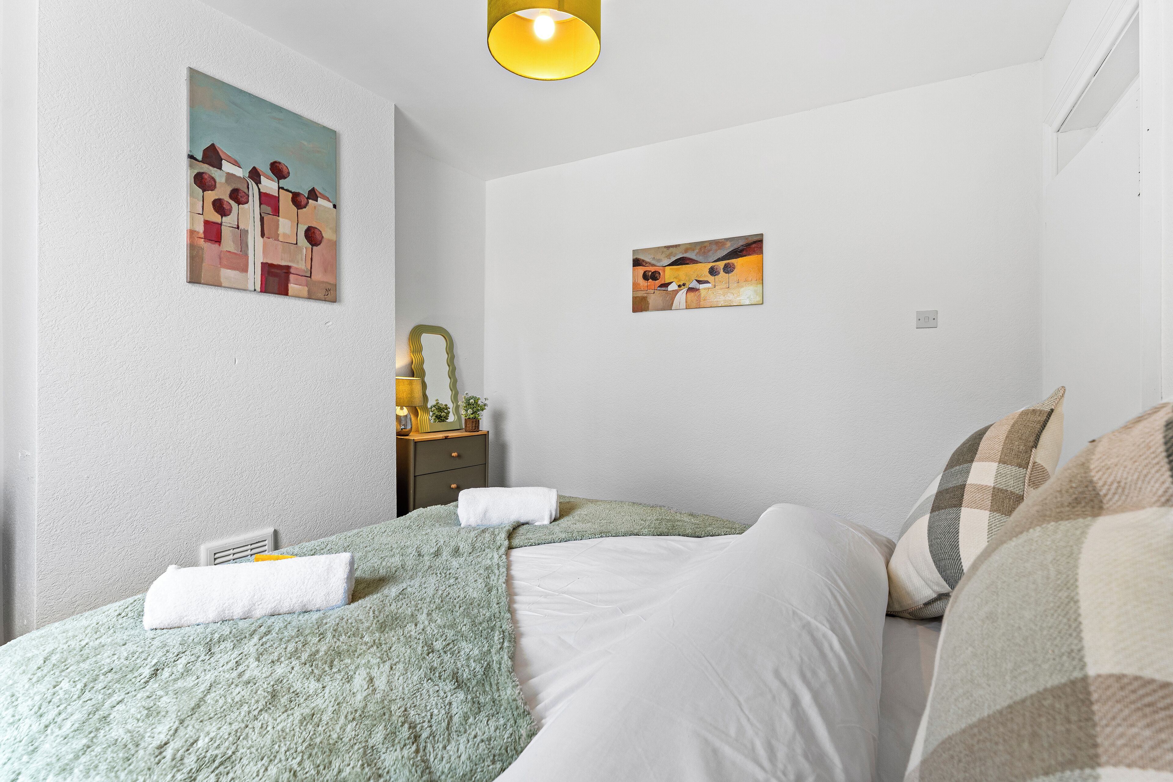 A bedroom with a bed, two paintings, a bedside table with a lamp, and a window.