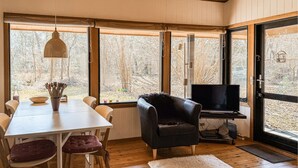 Ein gemütliches Zimmer mit Esstisch, Stühlen, einem schwarzen Ledersessel, einem Fernseher und großen Fenstern mit Blick auf Bäume.