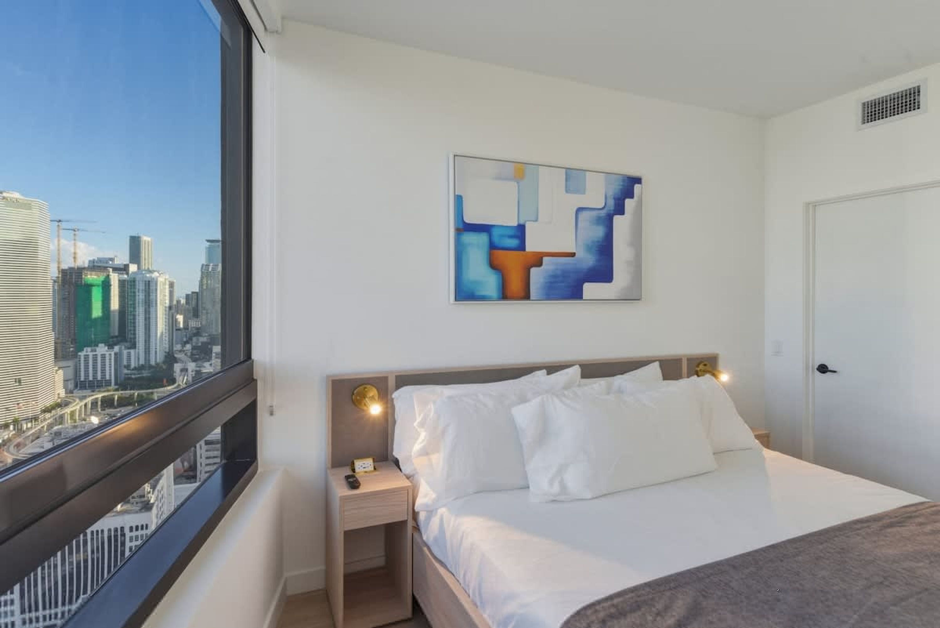 A hotel room with a city view, a bed with white bedding, a nightstand with a lamp, and a modern abstract painting on the wall.