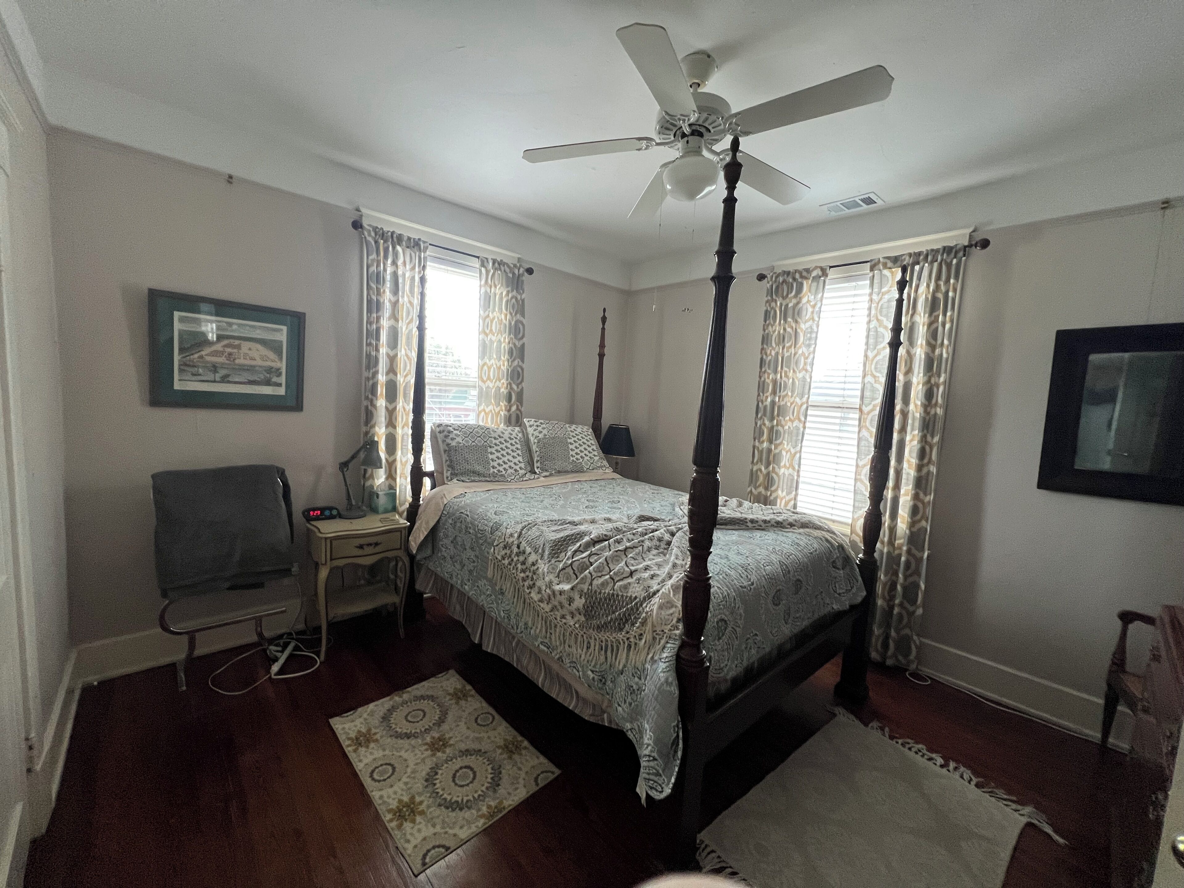 A bedroom with a four-poster bed, a ceiling fan, a bedside table with a lamp, a chair, and a rug.