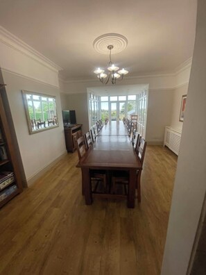 A spacious dining area with a long wooden table, chairs, a chandelier, and large windows.