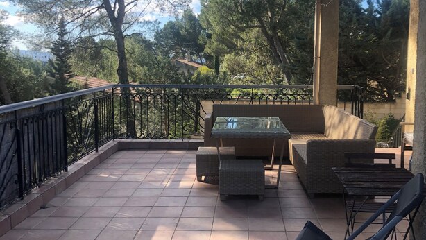 Eine überdachte Terrasse mit Gartenmöbeln, einem gefliesten Boden und Blick auf Bäume sowie ein Gebäude in der Ferne.