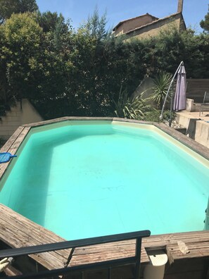 A rectangular pool with a wooden deck edge, surrounded by trees and a building in the background. - House surrounded by pine trees, 20 min from Avignon and St Rémy de Provence (Chateaurenard)