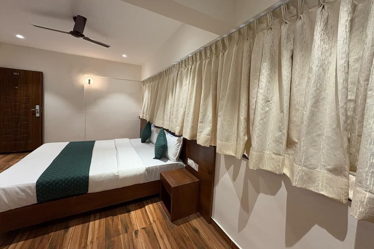 A bedroom with a wooden floor, a bed with a green and white bedspread, a wooden nightstand, a ceiling fan, and a window with curtains.