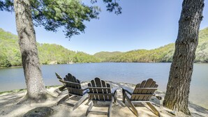 A lakeside view with three wooden Adirondack chairs under trees, a sandy beach, and a calm lake surrounded by forested hills. - 4 Mi to Bear Creek Lake! Serene Escape w/ Deck (Tuckasegee)