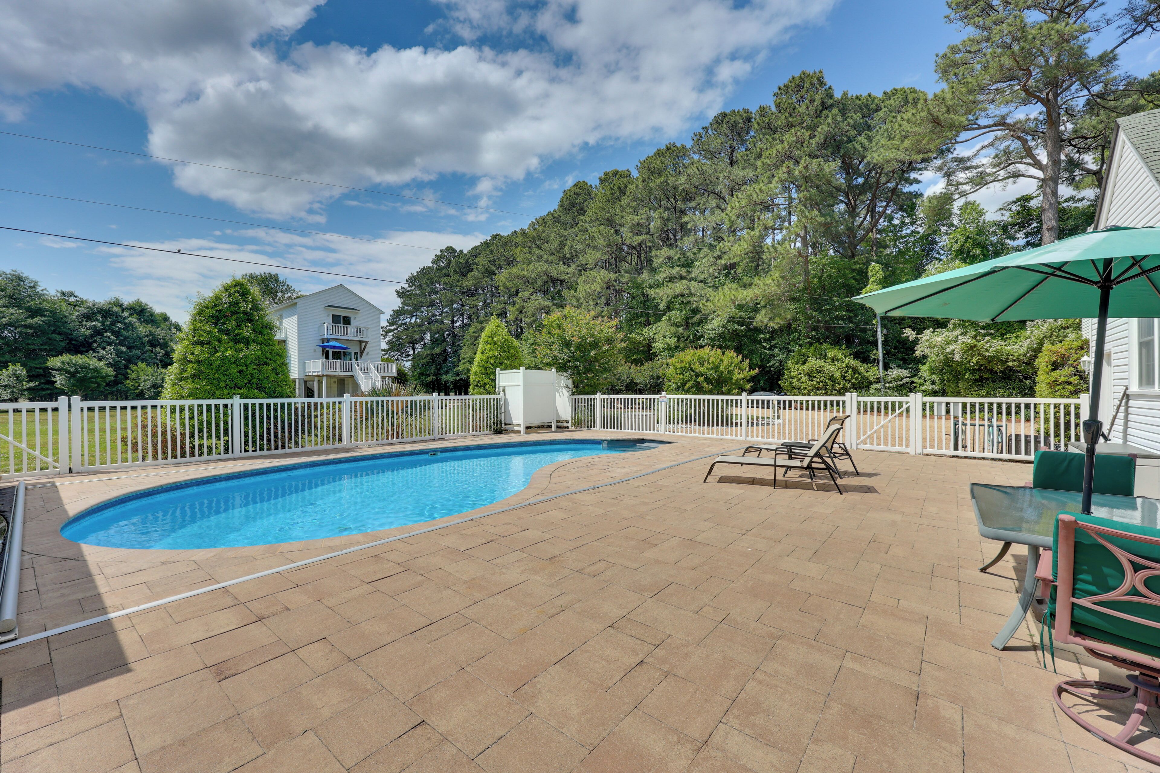 A swimming pool with a white fence, patio furniture, and a view of a house and trees.