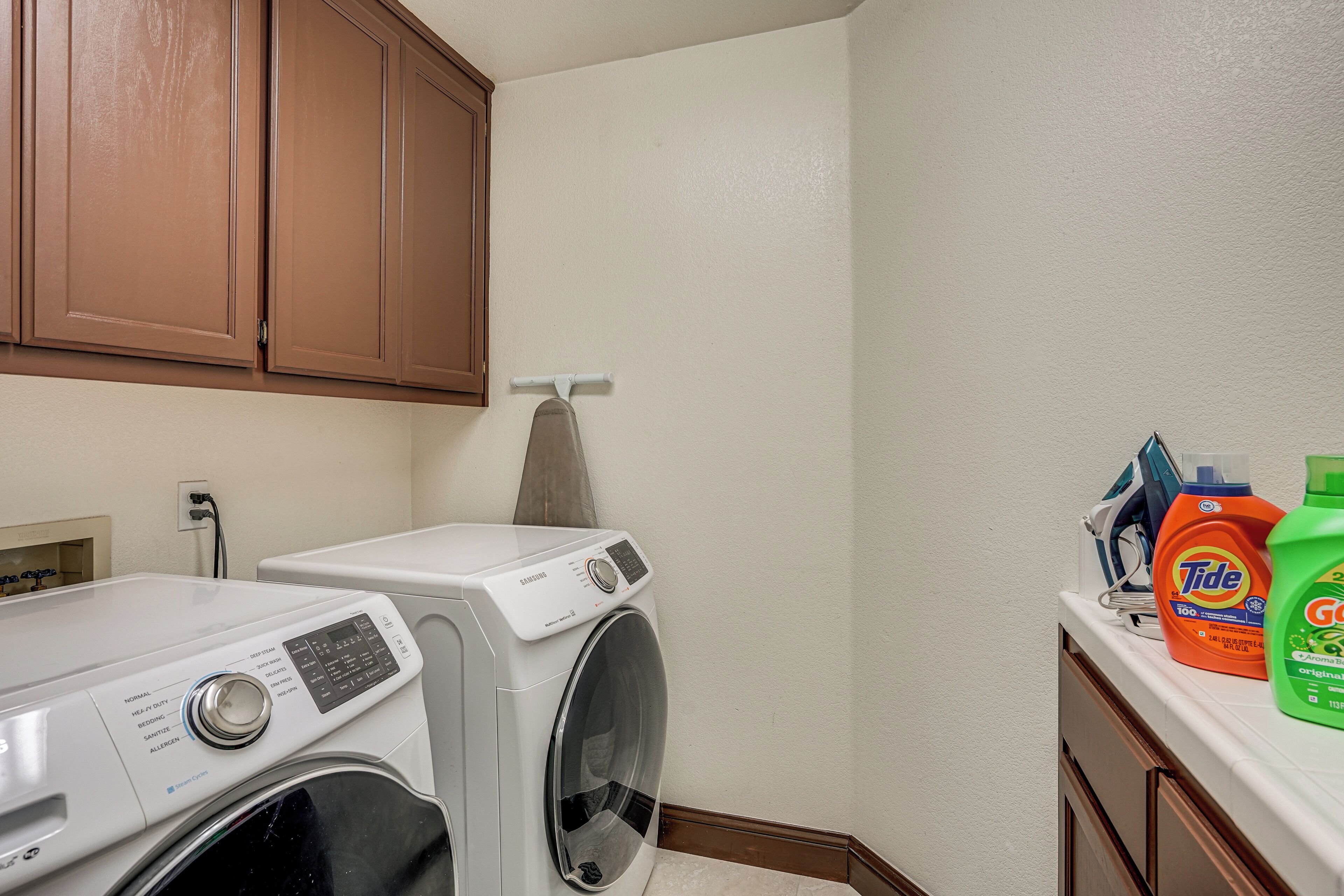 A laundry area with a front-loading washer and dryer, wooden cabinets, and Tide and Gain detergent bottles on a counter.
