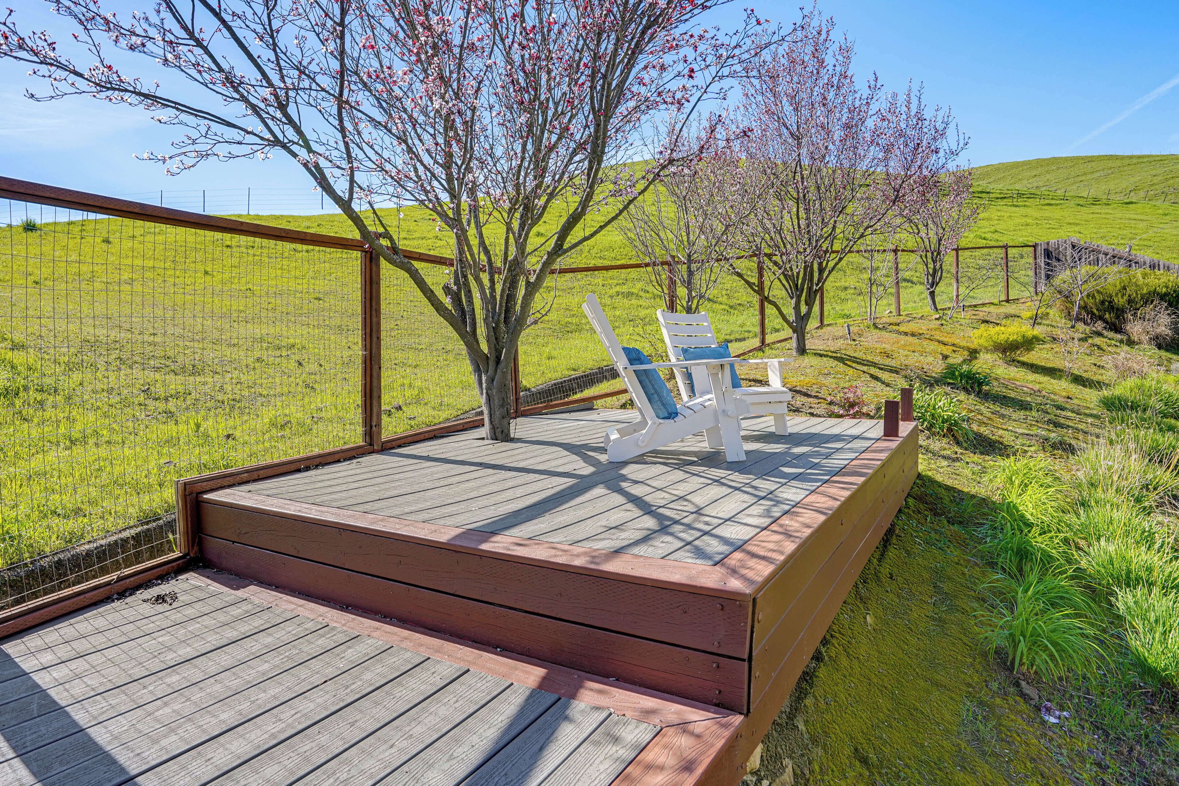 Una terraza de madera con dos sillas Adirondack blancas y una mesa, rodeada por una valla de madera y un campo cubierto de césped.