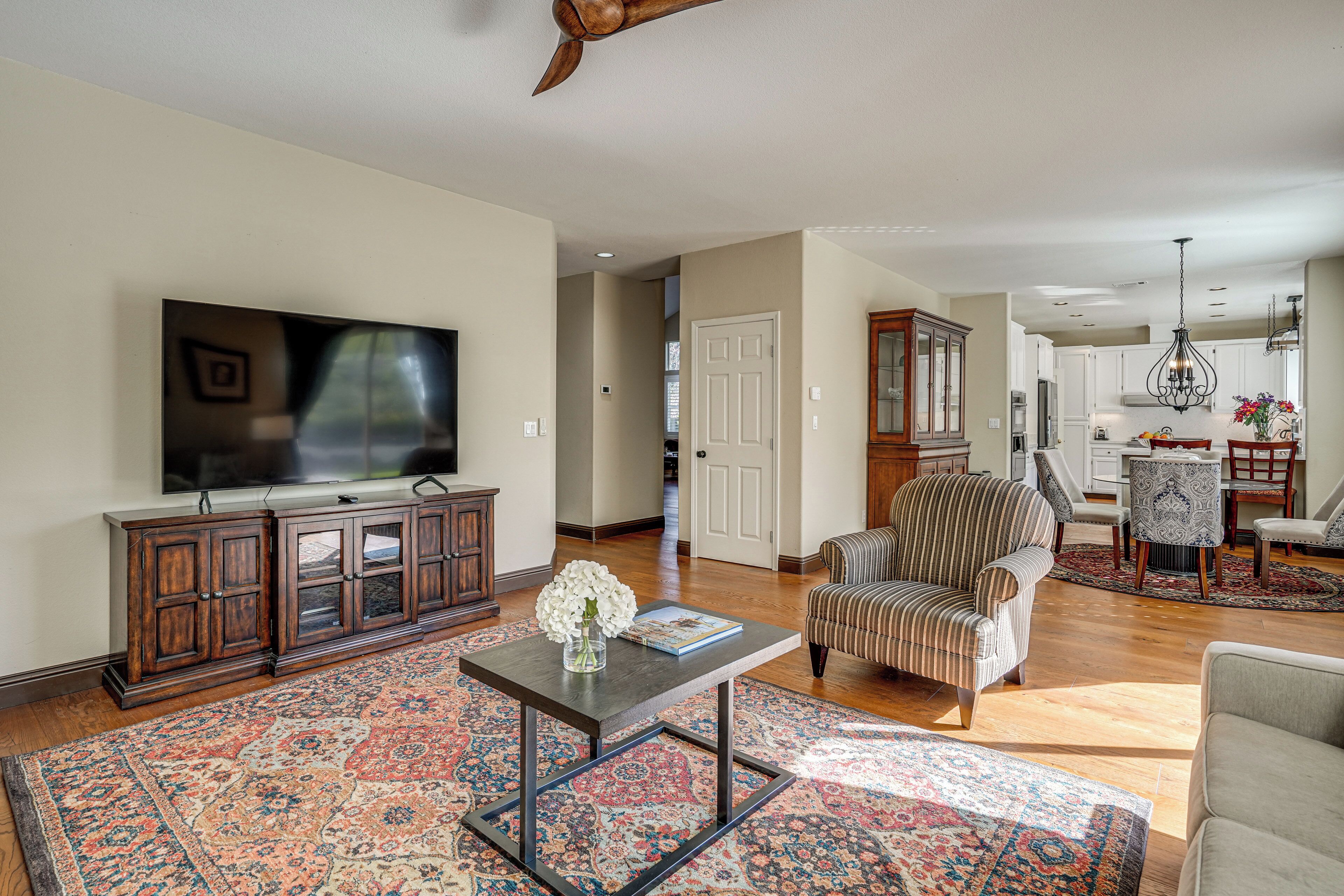 A spacious living room with a large flat-screen TV, a wooden entertainment center, a striped armchair, and a dining area with a chandelier.
