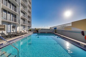 A swimming pool with a sunlit sky, a building with balconies, and lounge chairs. - Elegant Condo w/ Stunning Views in Dtwn Hollywood (Hollywood)