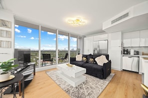 A modern living room with a black sofa, white coffee table, and a city view through large windows. - Elegant Condo w/ Stunning Views in Dtwn Hollywood (Hollywood)