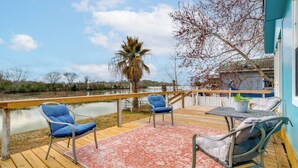 Um deck de madeira com mobiliário externo, vista para um corpo d'água e um edifício com fachada azul.
