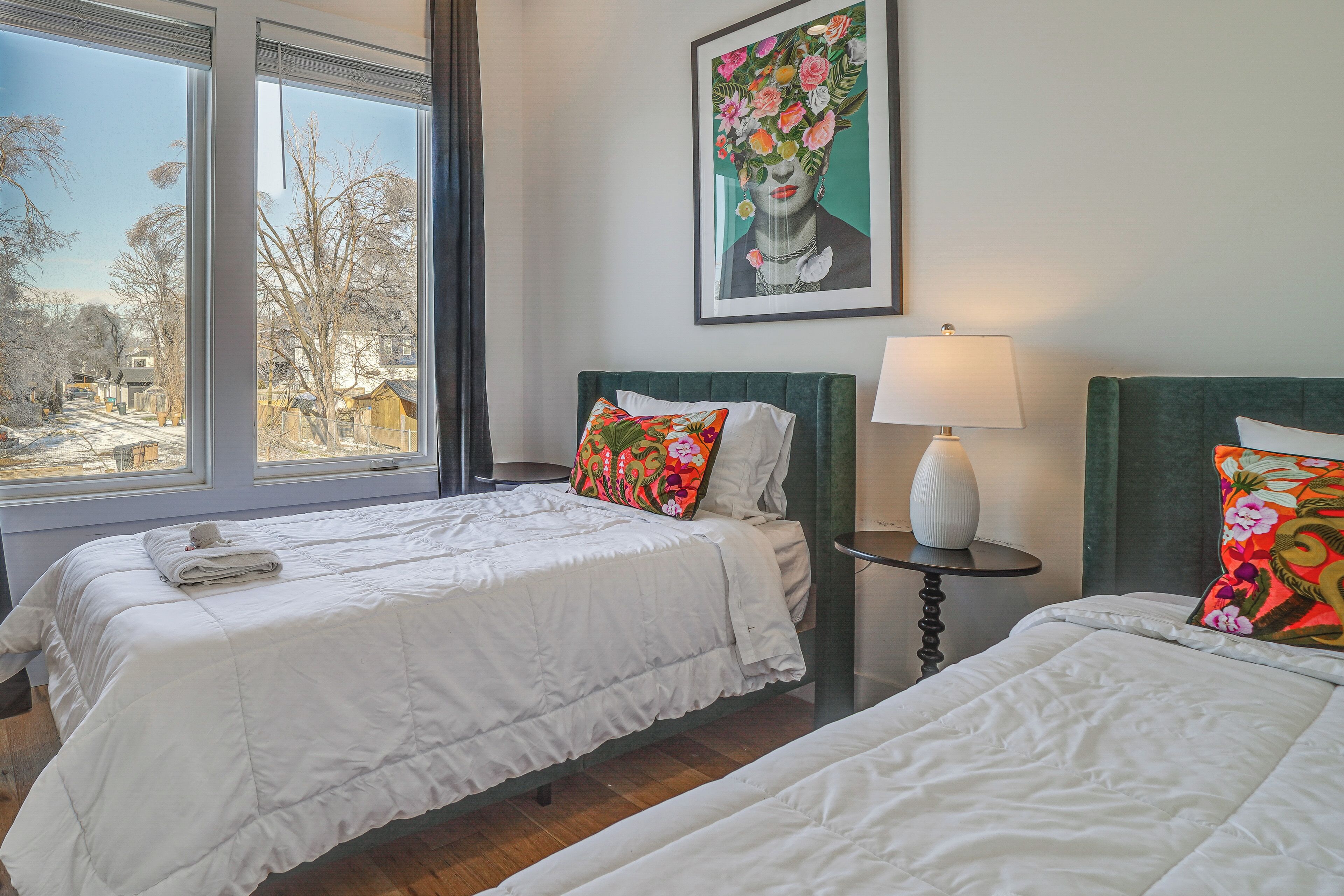 A hotel room with two beds, a window with a view of a snowy landscape, a painting on the wall, and a bedside table with a lamp.