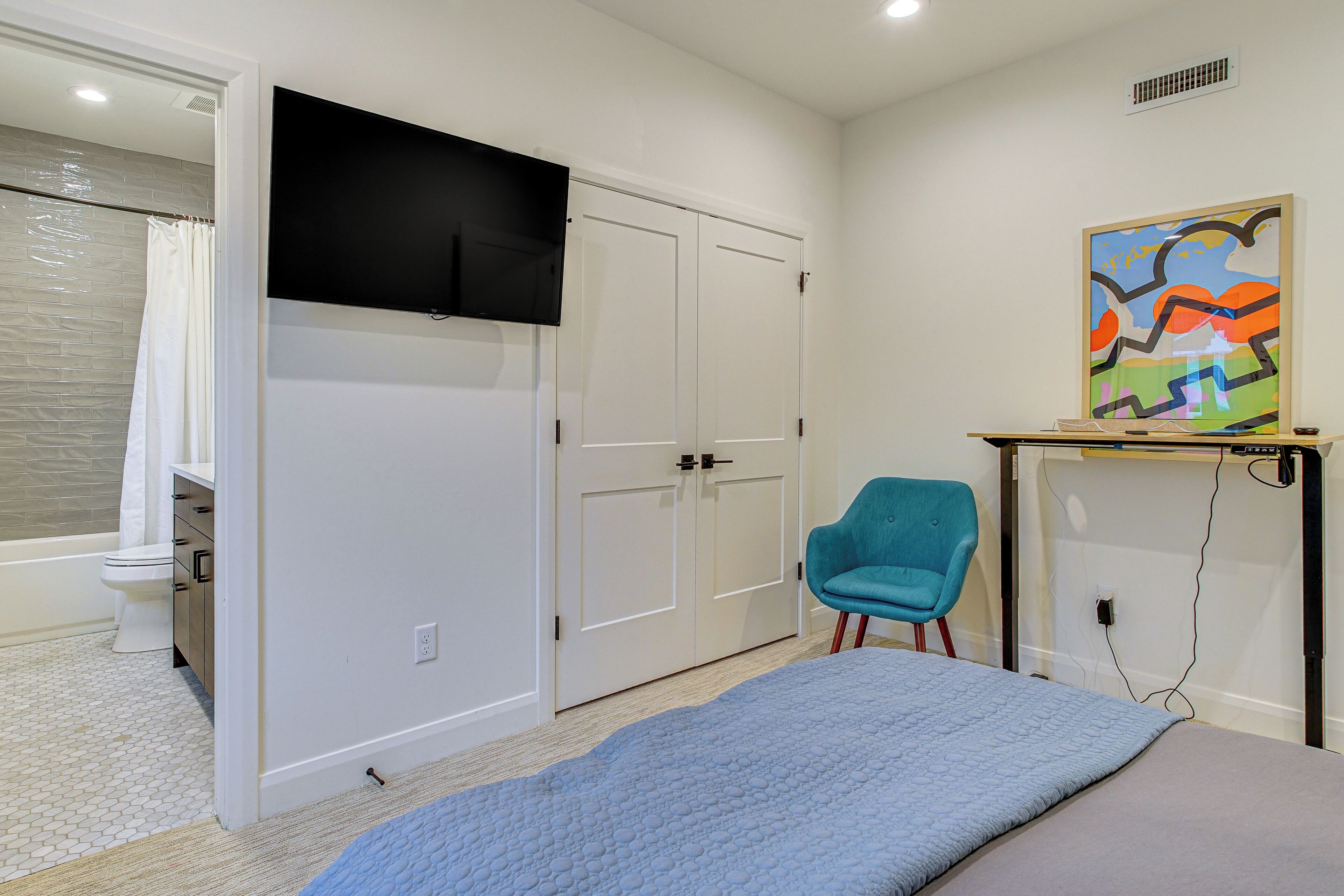 A bedroom with a bed, a chair, a TV, a painting, and a bathroom with a toilet and shower.